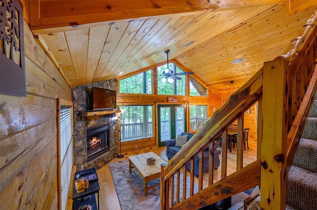 living room featuring wooden walls, wooden ceiling, ceiling fan, a stone fireplace, and stairway