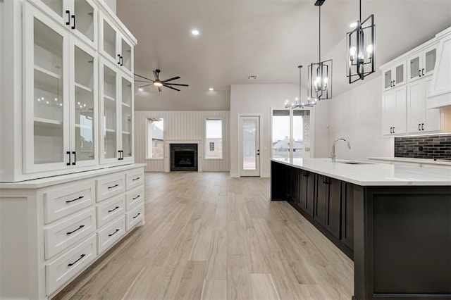 kitchen with a fireplace, a center island with sink, a sink, light wood finished floors, and ceiling fan with notable chandelier