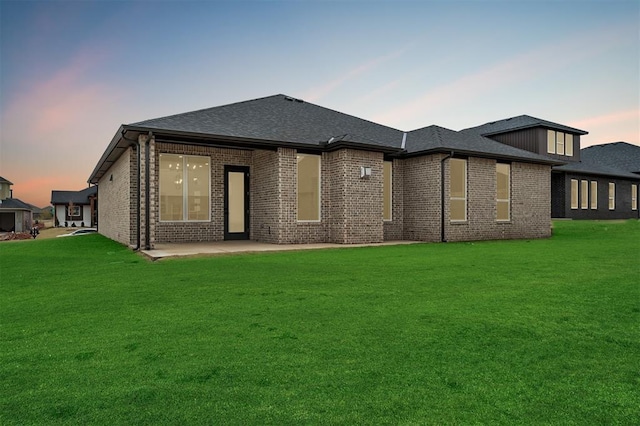 back of house at dusk featuring a patio, roof with shingles, a yard, and brick siding