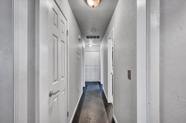 hallway featuring finished concrete floors, a textured wall, baseboards, and visible vents