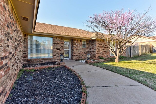 entrance to property with brick siding, visible vents, fence, and a lawn