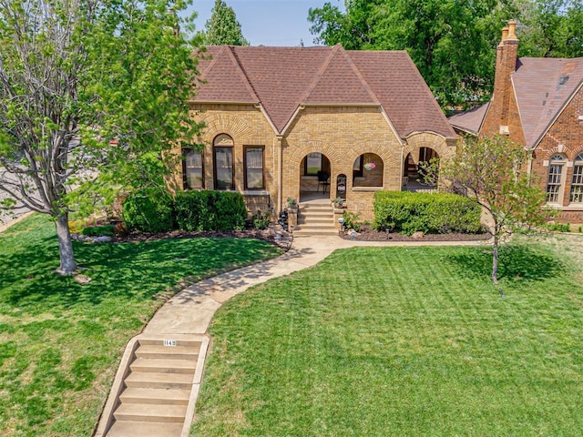 english style home with brick siding, a shingled roof, a front yard, and a chimney