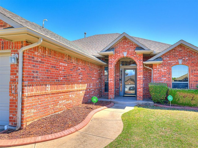 view of exterior entry featuring a yard, brick siding, and a shingled roof
