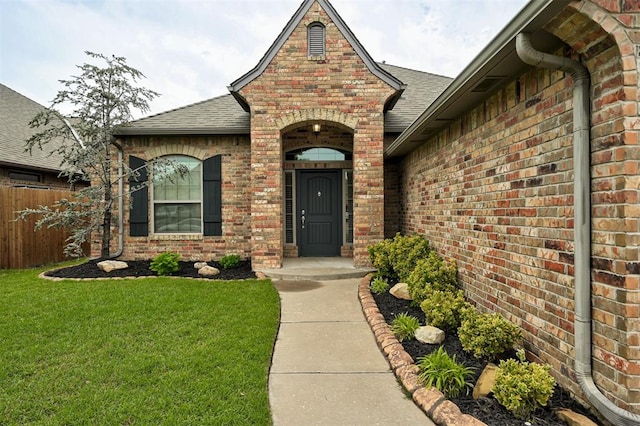 property entrance featuring fence, a lawn, a shingled roof, and brick siding