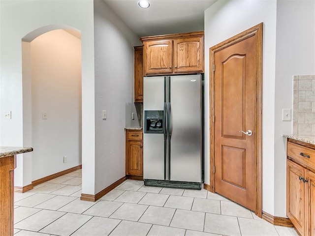 kitchen with light tile patterned flooring, light stone counters, stainless steel fridge with ice dispenser, arched walkways, and brown cabinetry