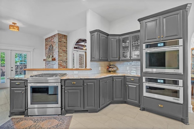 kitchen featuring gray cabinets, tasteful backsplash, stainless steel gas stove, light stone counters, and a peninsula