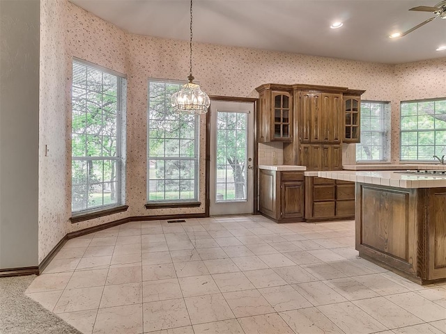 3601 Rena Dawn Ln, Edmond, OK 73013 - kitchen featuring tile countertops, hanging light fixtures, brown cabinets, glass insert cabinets, and wallpapered walls