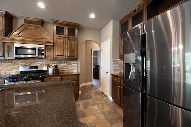 kitchen with baseboards, dark stone counters, tasteful backsplash, arched walkways, and stainless steel appliances