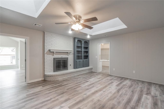 unfurnished living room with a ceiling fan, visible vents, a skylight, a brick fireplace, and wood finished floors