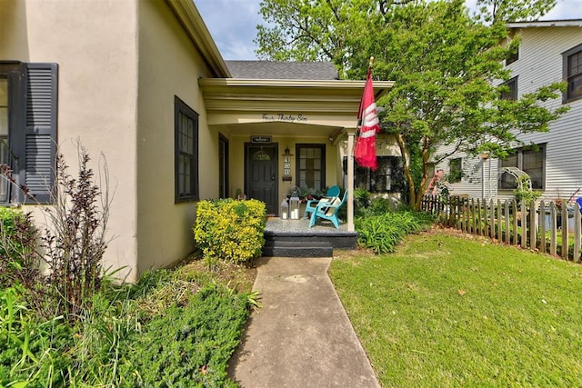 view of exterior entry with a lawn, fence, covered porch, stucco siding, and a shingled roof