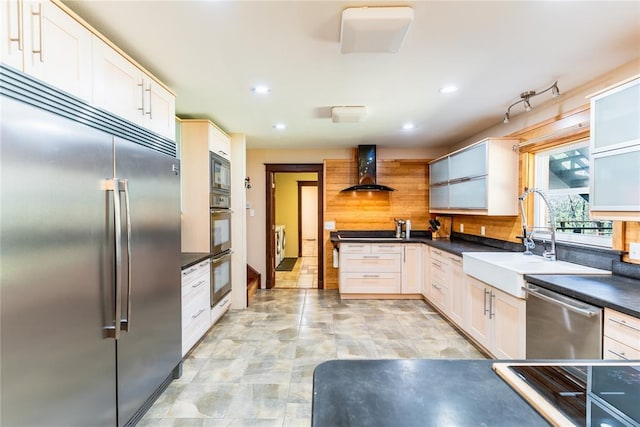 kitchen featuring wall chimney range hood, decorative backsplash, dark countertops, a sink, and built in appliances