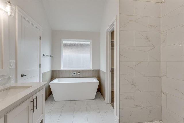 full bathroom featuring vanity, marble finish floor, wainscoting, a soaking tub, and vaulted ceiling