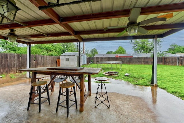 view of patio featuring a trampoline, ceiling fan, a shed, a fenced backyard, and an outbuilding