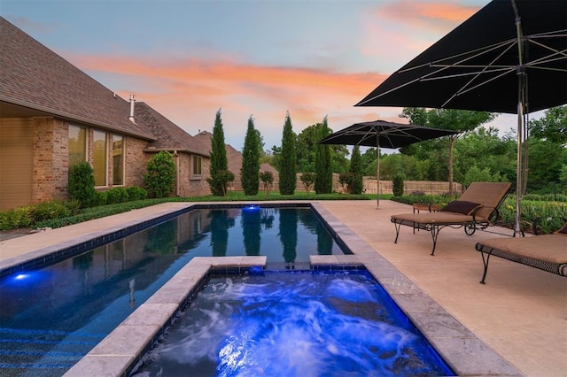 pool at dusk with fence, a pool with connected hot tub, and a patio area