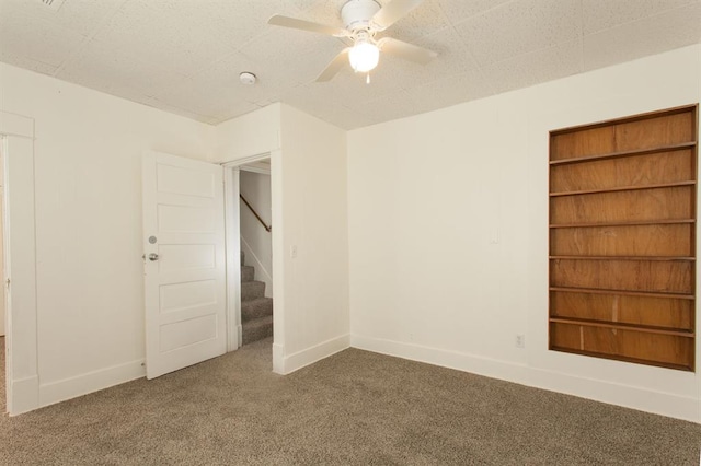 carpeted empty room with ceiling fan, built in features, baseboards, and stairway