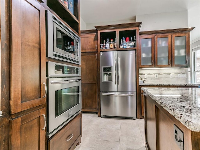 kitchen with backsplash, appliances with stainless steel finishes, light tile patterned floors, light stone counters, and glass insert cabinets
