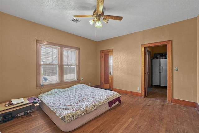 bedroom with a textured ceiling, hardwood / wood-style floors, freestanding refrigerator, baseboards, and visible vents