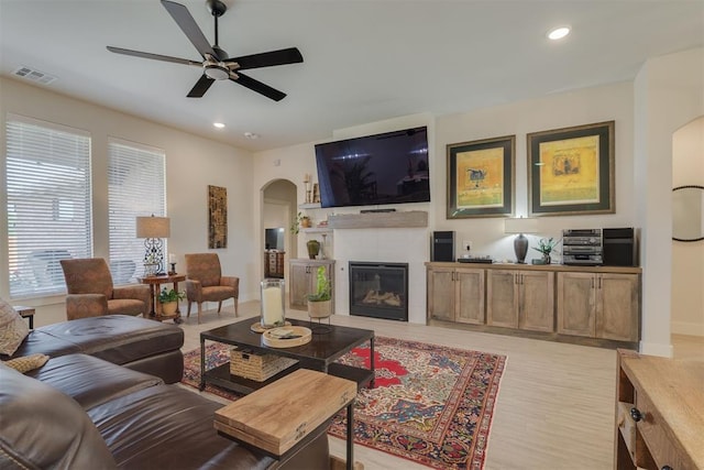 living room featuring a ceiling fan, visible vents, arched walkways, a tile fireplace, and light wood-style floors