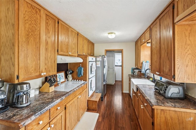 kitchen with white appliances, dark wood-type flooring, under cabinet range hood, brown cabinets, and a sink