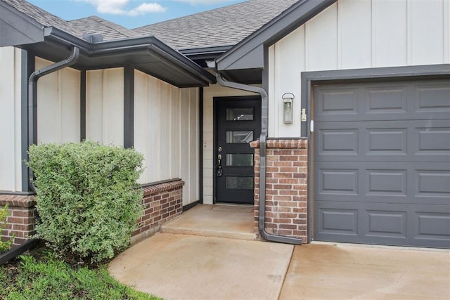 view of exterior entry with roof with shingles, brick siding, board and batten siding, an attached garage, and driveway