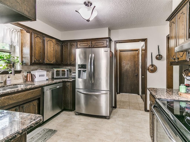kitchen with dark brown cabinetry, a sink, a textured ceiling, dark stone countertops, and stainless steel appliances