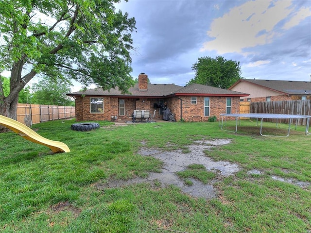 back of house featuring a fenced backyard, a chimney, brick siding, a lawn, and a trampoline