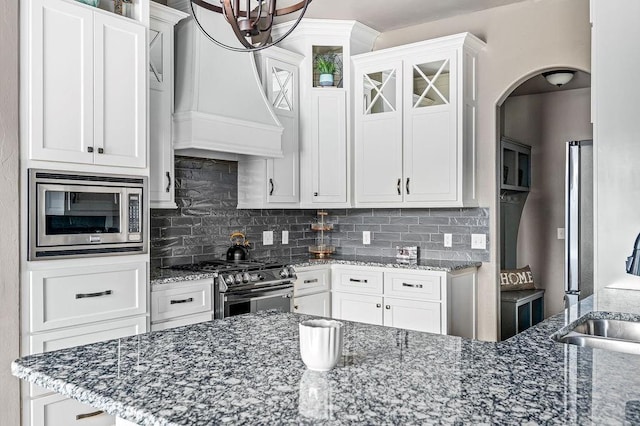 kitchen featuring dark stone counters, custom range hood, arched walkways, backsplash, and stainless steel appliances