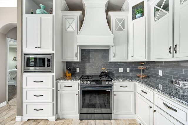 kitchen with dark stone countertops, stainless steel appliances, white cabinetry, backsplash, and custom exhaust hood