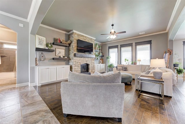 living room featuring crown molding, a stone fireplace, dark wood-style floors, arched walkways, and a ceiling fan