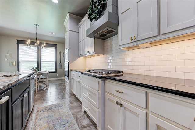kitchen featuring wall chimney range hood, decorative backsplash, appliances with stainless steel finishes, baseboards, and a notable chandelier