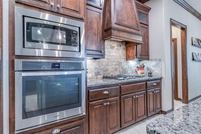 kitchen featuring decorative backsplash, custom exhaust hood, glass insert cabinets, light stone counters, and appliances with stainless steel finishes