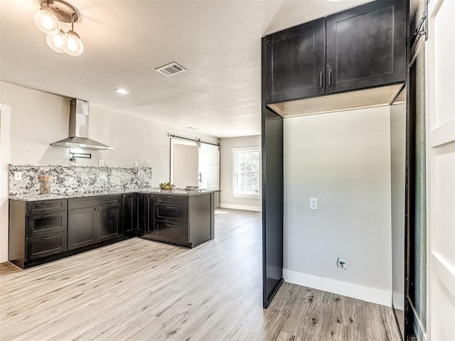 kitchen featuring dark cabinetry, wall chimney exhaust hood, a peninsula, light wood-type flooring, and visible vents