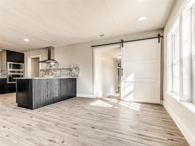kitchen featuring a barn door, light wood-style floors, wall chimney exhaust hood, stainless steel appliances, and visible vents