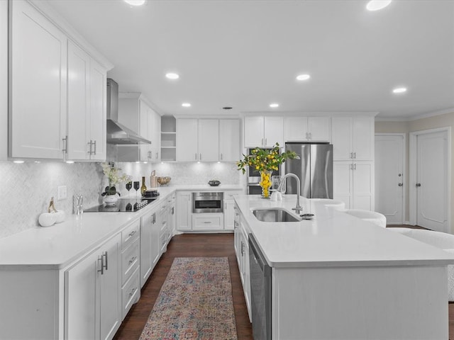 kitchen with wall chimney range hood, appliances with stainless steel finishes, a center island with sink, a sink, and white cabinetry
