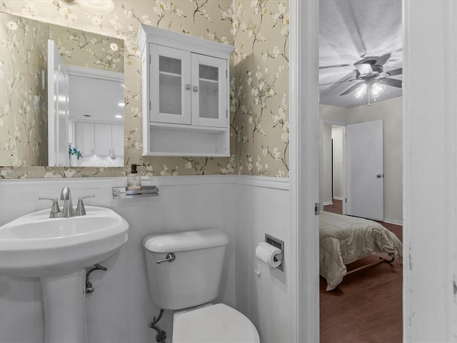 bathroom featuring wainscoting, toilet, a ceiling fan, wallpapered walls, and wood finished floors