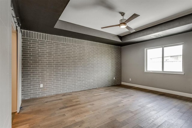 spare room with a ceiling fan, brick wall, wood finished floors, a raised ceiling, and baseboards