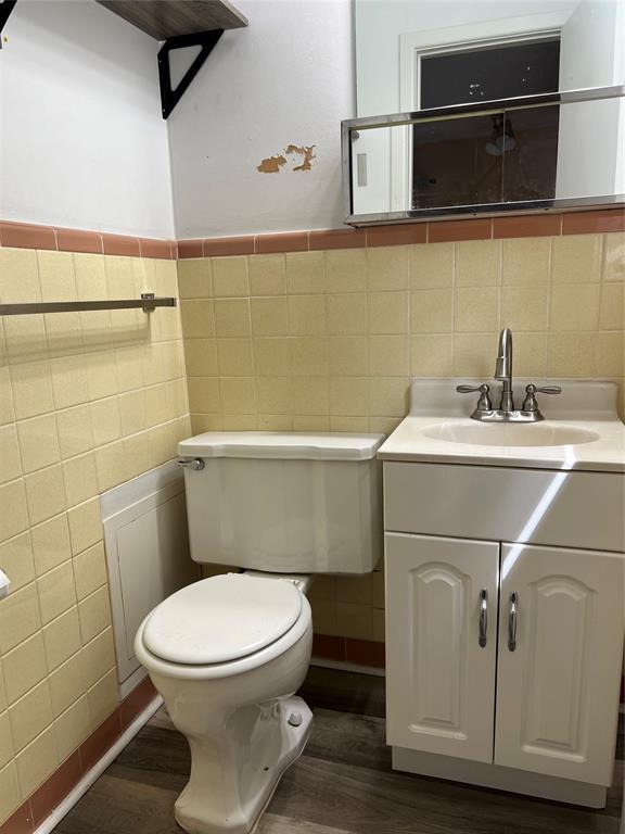 half bath featuring tile walls, wainscoting, wood finished floors, vanity, and toilet