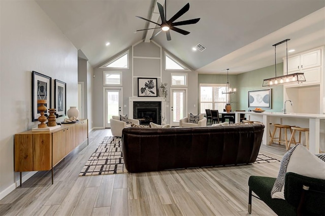 living area with a ceiling fan, a glass covered fireplace, high vaulted ceiling, light wood-style floors, and a chandelier