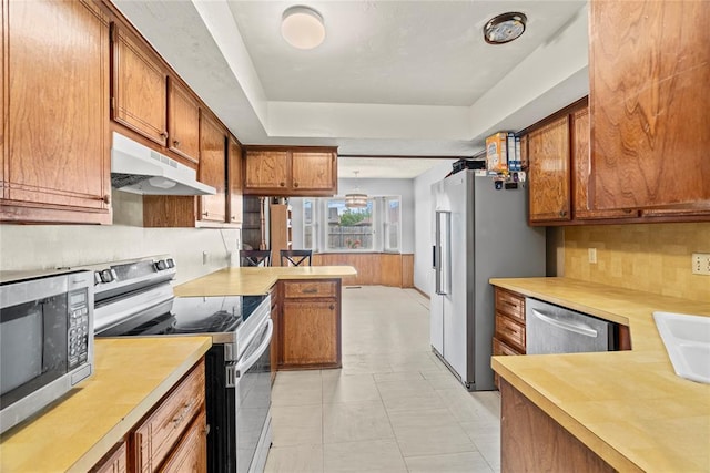 kitchen with light countertops, under cabinet range hood, a tray ceiling, brown cabinets, and appliances with stainless steel finishes