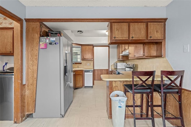 kitchen with brown cabinets, light countertops, a peninsula, under cabinet range hood, and stainless steel appliances