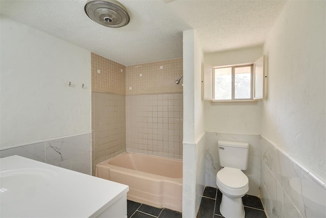 bathroom featuring toilet, tile patterned floors, a textured ceiling, a wainscoted wall, and tile walls