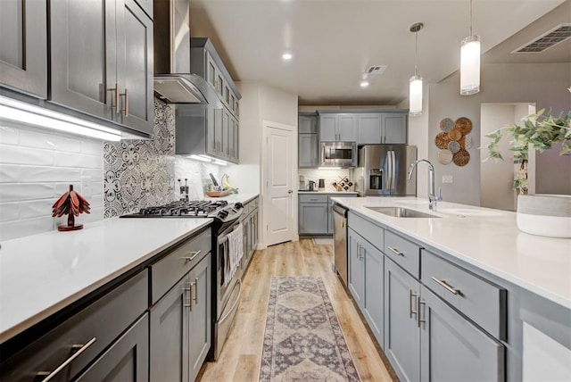 kitchen featuring wall chimney range hood, gray cabinets, a sink, stainless steel appliances, and light wood-style flooring