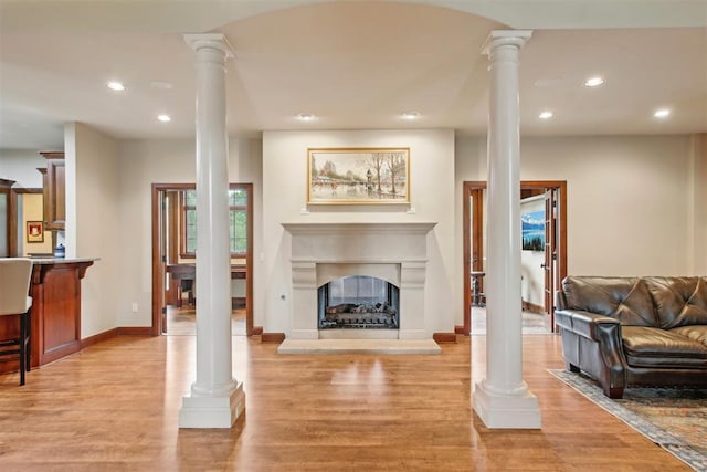 living area featuring baseboards, a fireplace with raised hearth, ornate columns, light wood-style floors, and recessed lighting