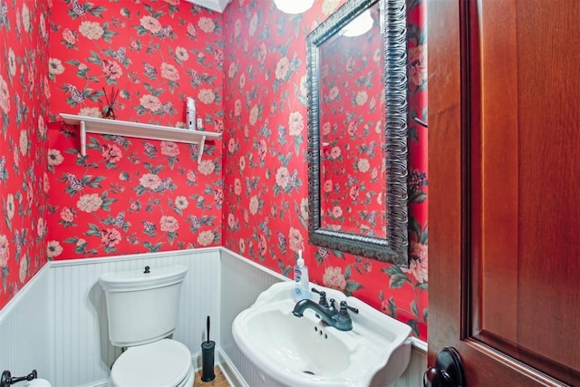 bathroom with toilet, wallpapered walls, wainscoting, and a sink