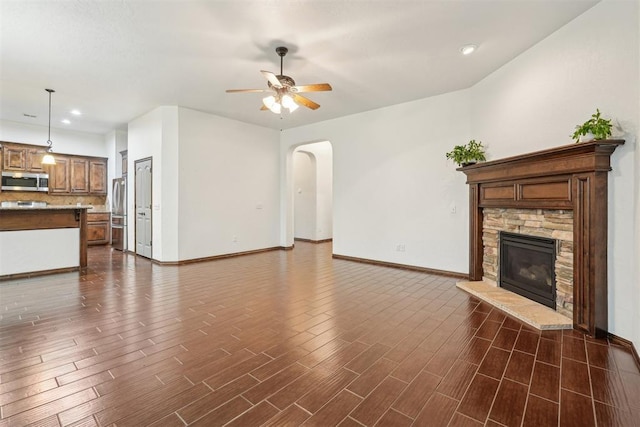 unfurnished living room with ceiling fan, a stone fireplace, arched walkways, baseboards, and dark wood-type flooring