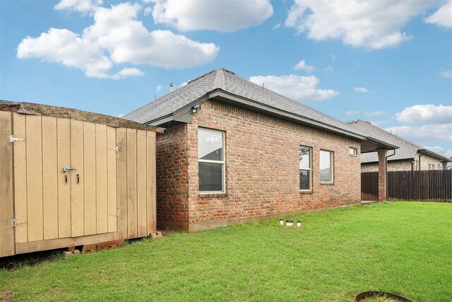 view of property exterior featuring an outdoor structure, fence, brick siding, a shingled roof, and a lawn