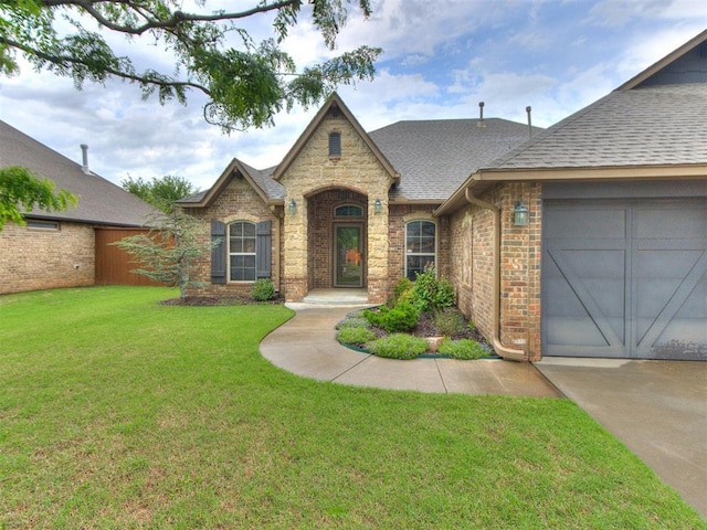 french country style house featuring a shingled roof, brick siding, a garage, stone siding, and a front yard