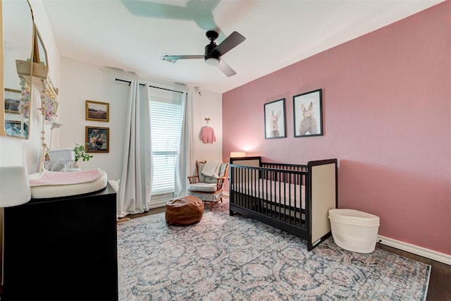 bedroom featuring baseboards, a nursery area, ceiling fan, and wood finished floors