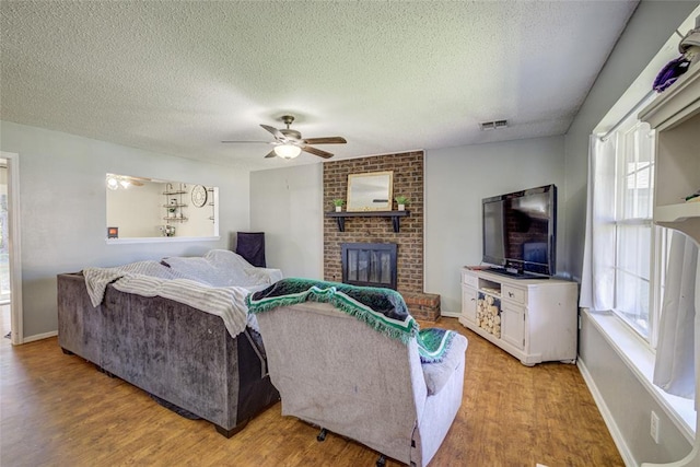 living room with ceiling fan, a textured ceiling, visible vents, a brick fireplace, and light wood-type flooring