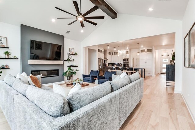 living area featuring visible vents, a glass covered fireplace, beam ceiling, light wood-style flooring, and ceiling fan with notable chandelier